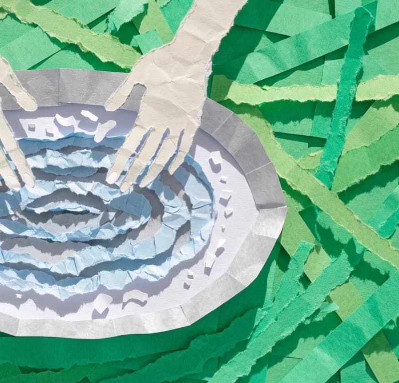 Cut-paper collage seen from above: torn paper shapes form a ring of blue water inside a silver rim on green paper grass, with two pale paper hands reaching in.